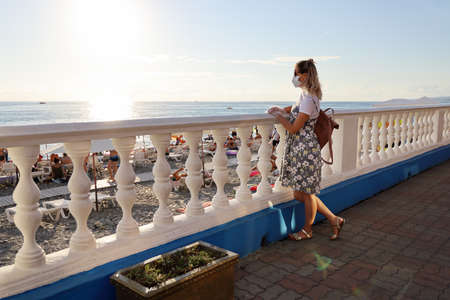 Young woman in protective mask stands alone near railing on waterfront and looks at beach, where people are resting. Sunset with copy spaceの写真素材