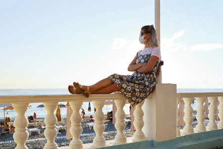Young woman in dress and protective mask on face, sitting alone on railing of promenade and looks at beach, where people restの写真素材