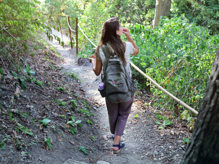 One young woman with backpack goes leisurely on forest path in summer sunny day