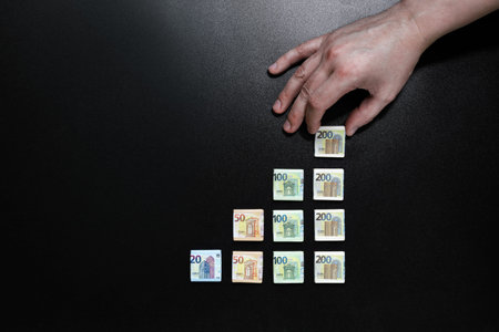 Profit concept, hand puts Euro banknote at top of steps of financial success. Over black background...