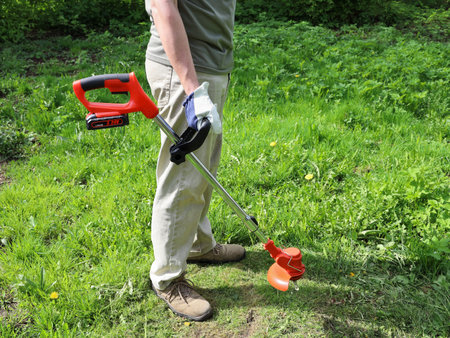 Man stands on meadow and holds an electric trimmer for mow grass, on sunny day in summer