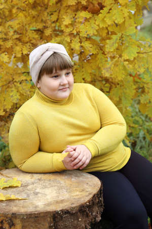 Obese smiling girl sits on a wooden stump near autumn trees with yellow foliageの写真素材