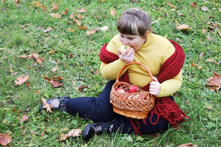 Obese girl with red scarf sits on the grass and holds basket with apples in autumnの写真素材