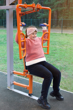 Chubby girl performs sports exercises on street simulators outdoors