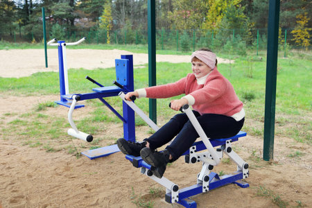 Chubby joyful girl performs sports exercises on street simulators outdoors