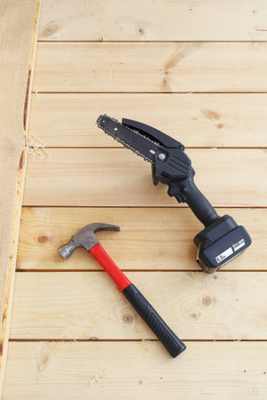 Electric chain saw with battery for wood sawing, small work, on wooden surface next to hammer