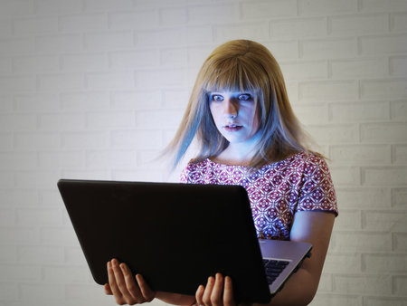 Girl teenager with blond hair widely open her eyes from screen laptop in with blue light, against...