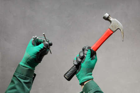 Hands in worn working gloves hold nails and hammer, close-up on textured gray backgroundの写真素材