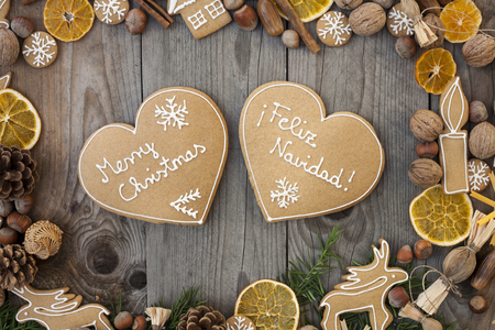 Two heart shaped gingerbreads with white icing, on wooden background, with Merry Christmas written in English and Spanishの写真素材