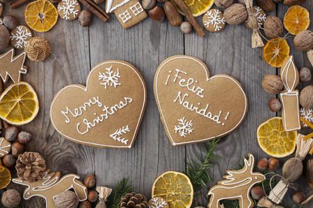 Two heart shaped gingerbreads with white icing, on wooden background, with Merry Christmas written in English and Spanishの写真素材
