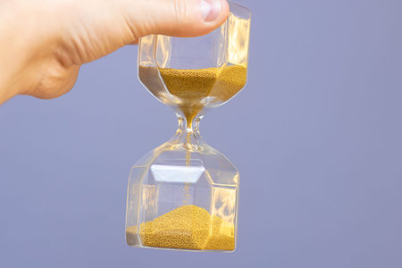 Hand holds transparent glass hourglass with falling yellow-gold sand closeup on blue wall background