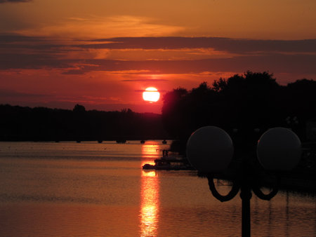 A large red sun sinks over the horizon against the backdrop of a street lamp and a lakeの写真素材