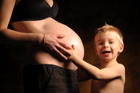Pregnant woman wearing a black bra and a little blond smiling boy touching her belly