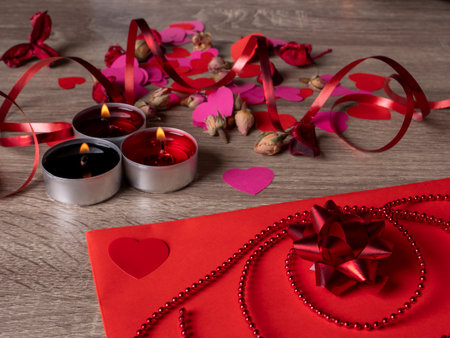 red envelope next to candles with red rose petals and ribbon on the tableの写真素材