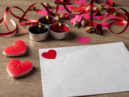 red envelope next to candles with red rose petals and ribbon on the tableの写真素材