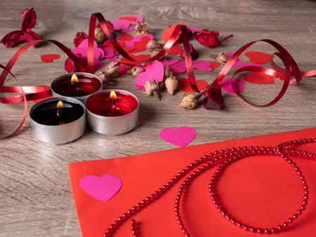 red envelope next to candles with red rose petals and ribbon on the tableの写真素材