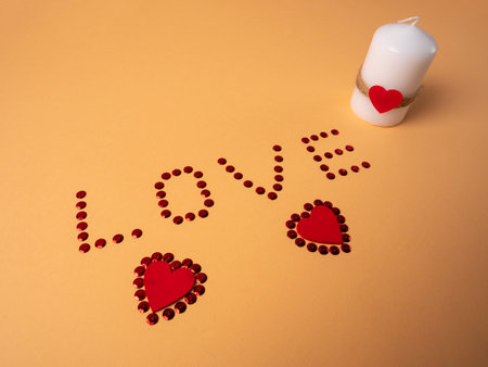 Sequins in the form of the inscription love and below two hearts that lie in sequins and next to a white candle on an orange backgroundの写真素材