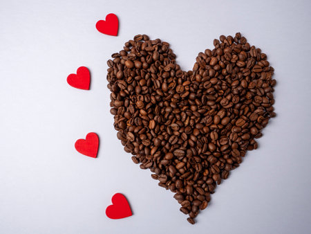 A large brown heart made from coffee beans lies next to four red hearts on a bright white backgroundの写真素材