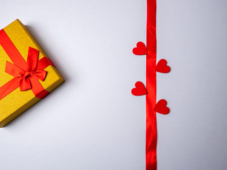On a white bright background lies a long red ribbon surrounded by four hearts and next to a yellow gift with a red ribbonの写真素材