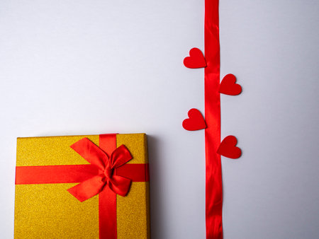 On a white bright background lies a long red ribbon surrounded by four hearts and next to a yellow gift with a red ribbonの写真素材