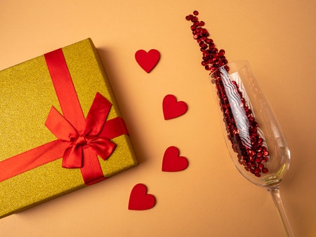 A yellow gift with a ribbon in the form of a butterfly knot lies next to four red hearts and a glass with red sparkles on an orange backgroundの写真素材