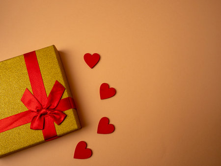A yellow gift with a ribbon in the form of a butterfly knot lies next to four red hearts on a nice orange backgroundの写真素材