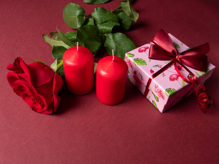 A large red rose that is next to two red large candles and a pink gift that are on a red backgroundの写真素材