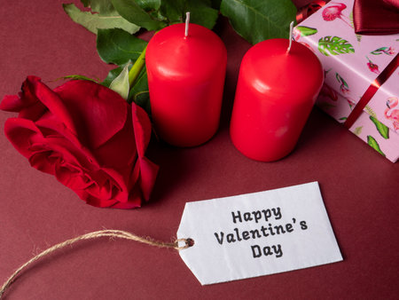 A large red rose that is next to two red large candles and a pink gift and a white note that are on a red backgroundの写真素材