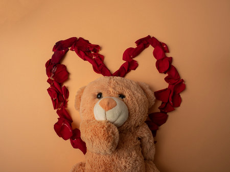 Many red rose petals lie in the shape of a large red heart and in the center of this heart lies a teddy bear on an orange backgroundの写真素材