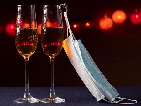 Two glasses with champagne, one glass weighs a ring and a blue medical mask on the ring on a black background with lightの写真素材