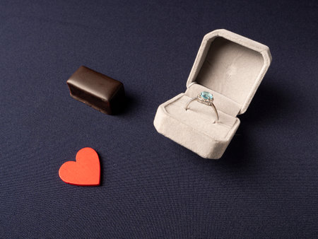 White box in which a ring with a blue stone, a red heart and chocolate lies nearby on a blue backgroundの写真素材