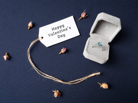 White box in which there is a ring with a blue stone and next to it lies a white note in which there is an inscription and around dry rose buds on a blue backgroundの写真素材