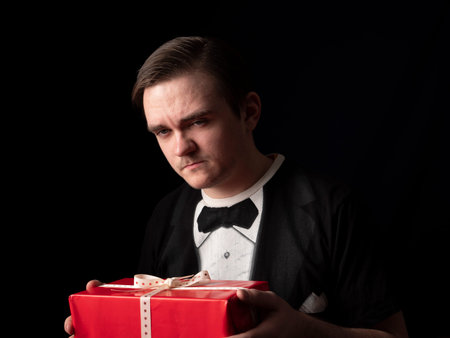 a young guy in a black t-shirt suit holds a red gift in his hands and thinks on a black background