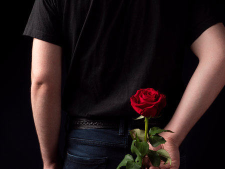 Young guy in a black t-shirt suit holds a red rose behind him on a black backgroundの写真素材