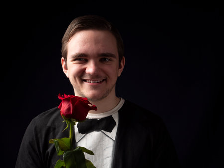 Young guy in a black T-shirt suit holds a red rose in his hands and smiles on a black backgroundの写真素材