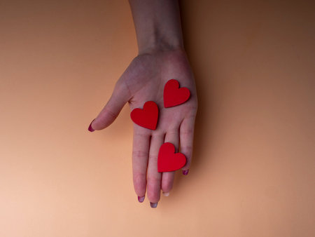 Stylish female manicure. Young female hands hold three red hearts, nails with red manicure, on an orange background. Manicure at home during a pandemicの写真素材