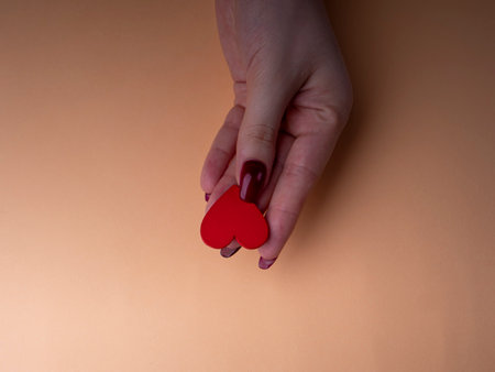 Stylish female manicure. Young female hand holds one red heart, nails with red manicure, on an orange background. Manicure at home during a pandemicの写真素材