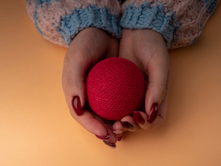 Stylish female manicure. Young female hands in a woolen sweater hold a red ball. nails with red manicure and white with hearts, on an orange background. Manicure at home during a pandemicの写真素材