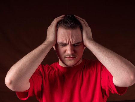 A young guy in a red T-shirt holds his head with his hands and thinks sadly against an orange...