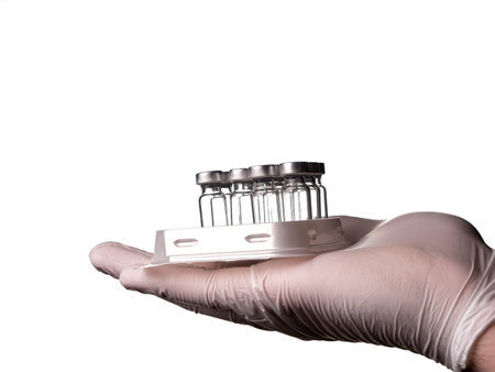 A doctor in white medical gloves holds glass vials with vaccine liquid in the tray of an automatic liquid dispenser. Glass vials and hand isolated on white backgroundの写真素材