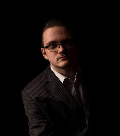 A portrait of a 20-30 year old young business man sits in glasses, a black suit and a white shirt and looks with a serious look. Young business man isolated on black backgroundの写真素材