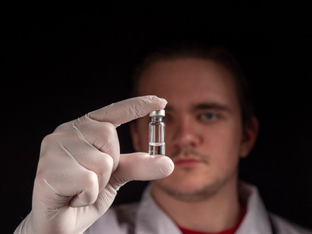 A young doctor holds a glass vial in which the liquid is with a vaccine in his outstretched hand. Selective focus on glass vial isolated on black backgroundの写真素材