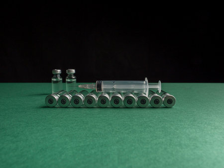 Glass vials in which the liquid with the vaccine are in a row and behind them there are two vials that stand and a syringe lies next to them. Glass vials are on a green and black backgroundの写真素材