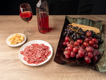 Sliced ââsalami and cheese with red wine, two type grapes, and Italian grissini on a tray, and on a wooden table, over black backgroundの写真素材