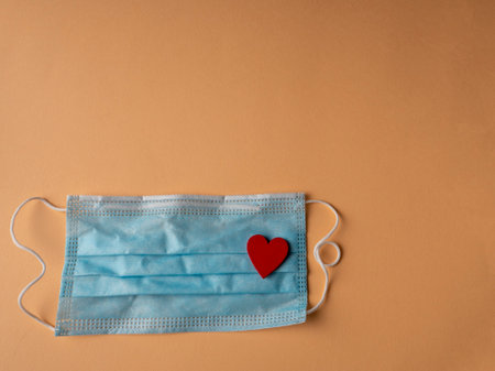 Blue medical mask on which lies the shape of a red heart on an orange backgroundの写真素材
