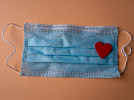 Blue medical mask on which lies the shape of a red heart on an orange backgroundの写真素材