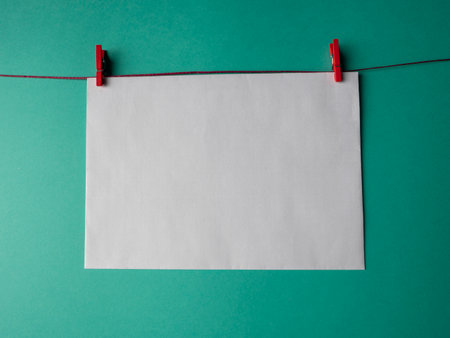 White paper that weighs on a red rope and is secured with two clothespins on a green background. A template for your project for valentine's dayの写真素材