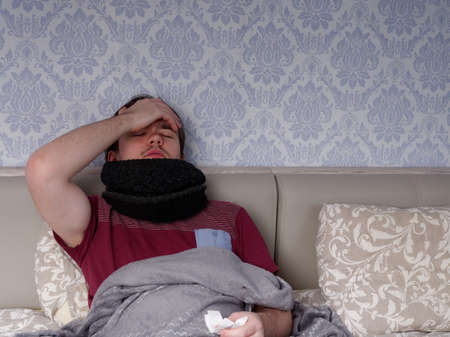 young guy in a red t-shirt sits on the bed and takes cough pills, pain relievers and cold medications, the concept of illness at homeの写真素材