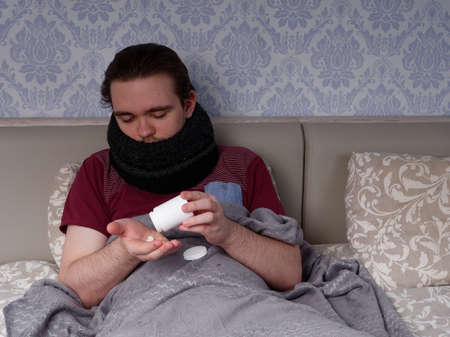 young guy in a red t-shirt sits on the bed and takes cough pills, pain relievers and cold medications, the concept of illness at homeの写真素材
