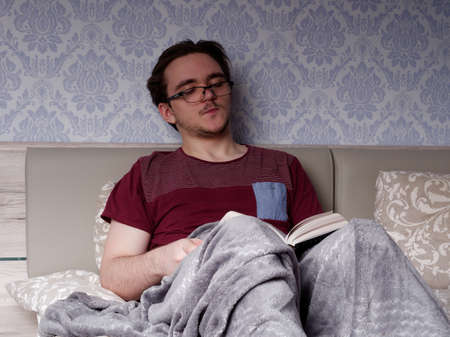 a young guy in a red T-shirt sits on a light-colored bed and reads a thick book, the concept of rest after a working dayの写真素材